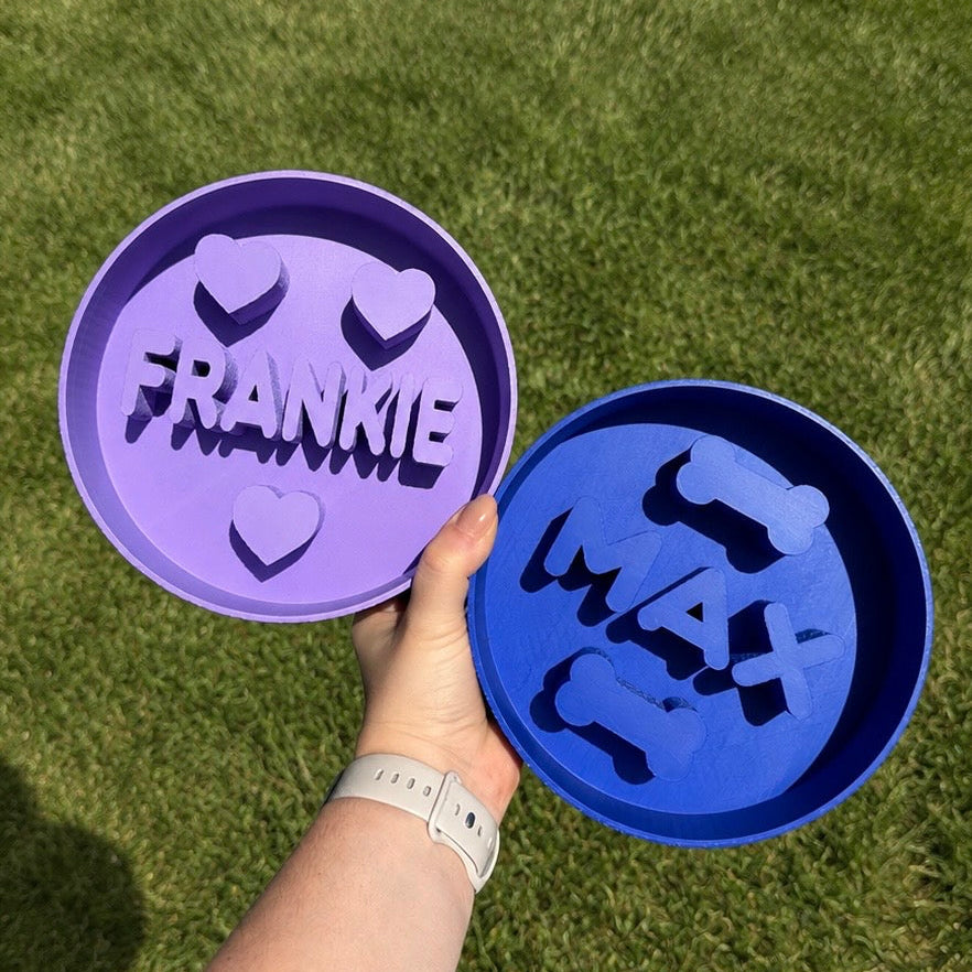 Two personalized pet bowls, one purple and one blue, held by a hand on grass.