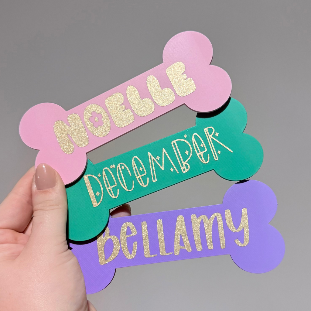 Hand holding three colorful bone-shaped name tags with glittery text on a gray background